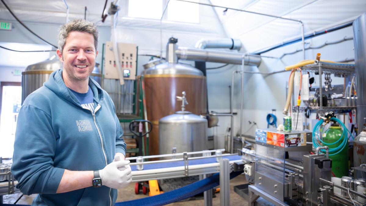 身穿蓝色运动衫的男子站在用于酿造啤酒的不锈钢设备前.